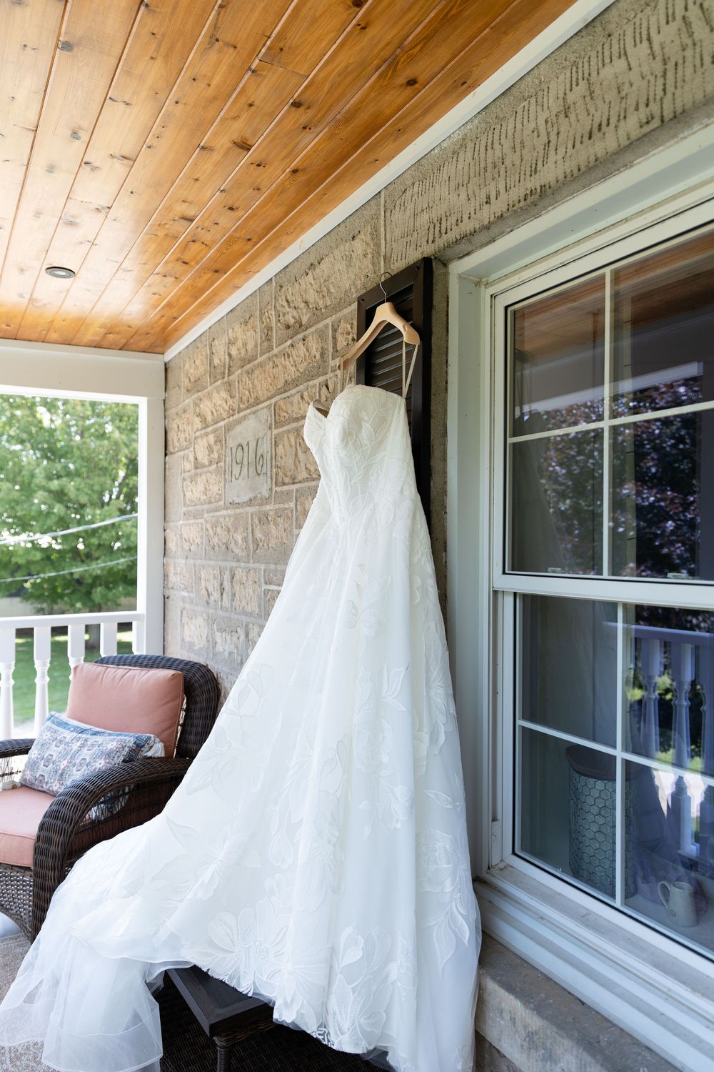 White strapless wedding dress hanging on a wooden hanger on a covered porch with wicker chairs on the morning of a Guelph, Ontario wedding.