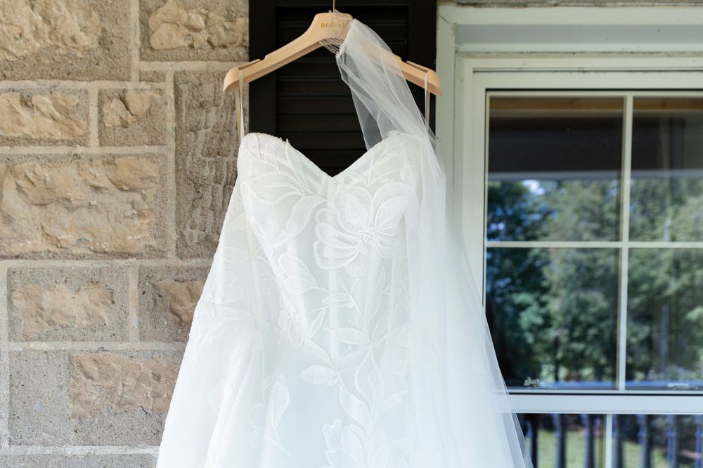 White strapless lace wedding dress with veil hanging on wooden hanger outside stone building on the morning of a wedding in Guelph, Ontario