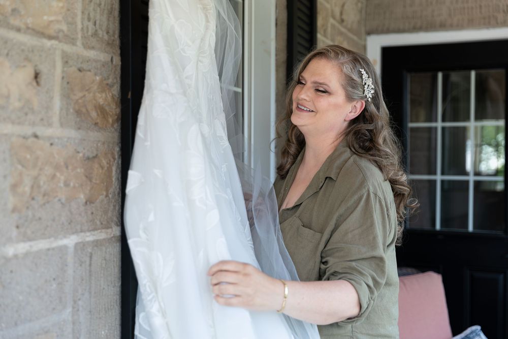 Bride admiring her white wedding dress hanging outdoors near stone wall on the morning of her wedding in Guelph, Ontario.