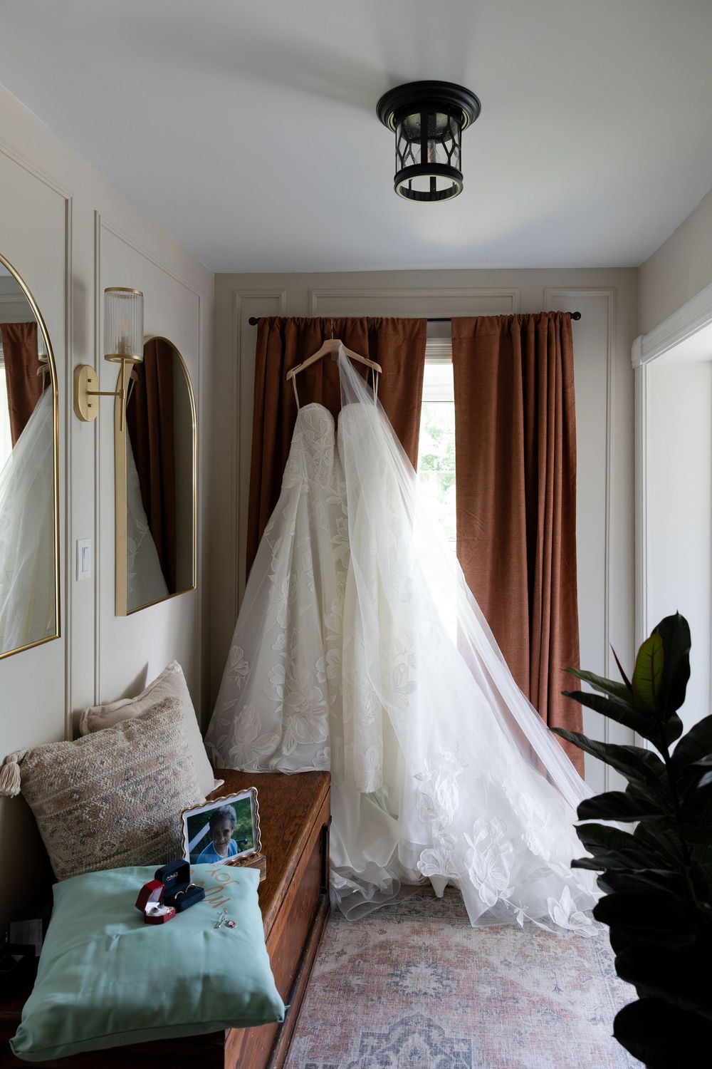 White lace wedding dress hanging on a hanger in an elegant room with rust curtains and a decorative bench in Guelph, Ontario.