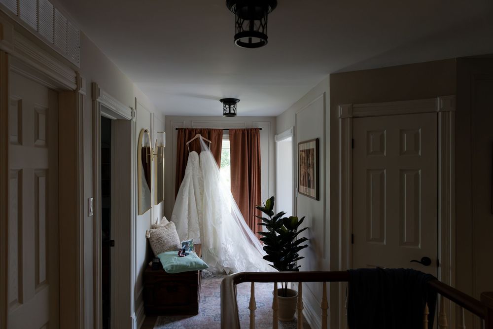 A white wedding dress hanging in a dimly lit hallway with warm curtains and a potted plant nearby in Guelph, Ontario.