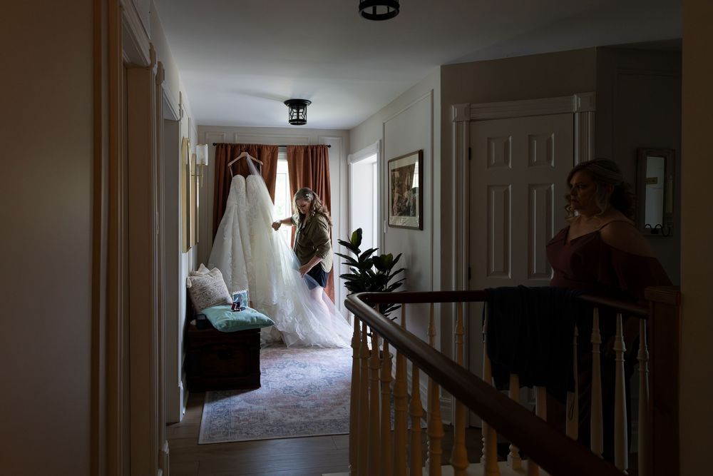 Bride holding her elegant white wedding gown in a warmly lit home hallway while a guest watches from the staircase in Guelph, Ontario.