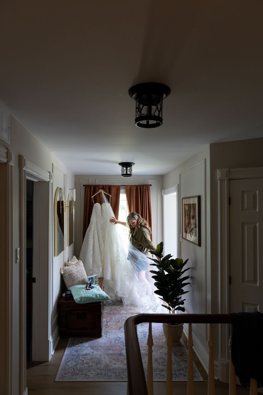 Bride holding her wedding gown in a softly lit hallway with warm decor, red curtains, and a fiddle leaf fig plant nearby in Guelph, Ontario.