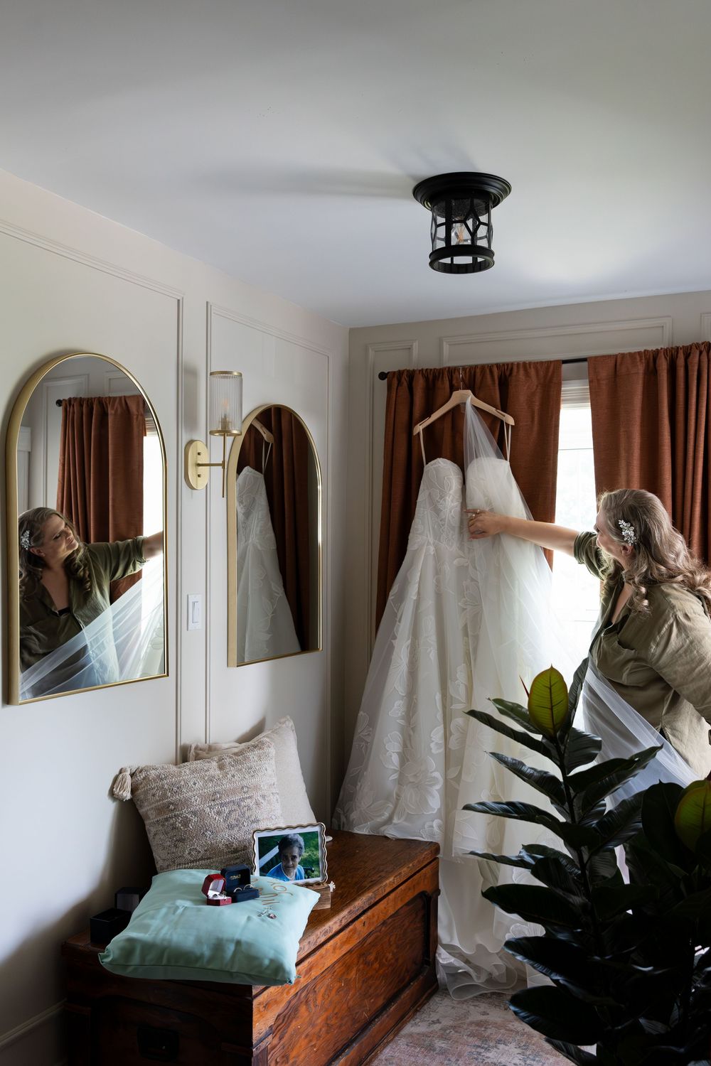 Woman holding a white lace wedding dress on a hanger near a window with rust curtains in an elegant bridal suite in Guelph, Ontario.
