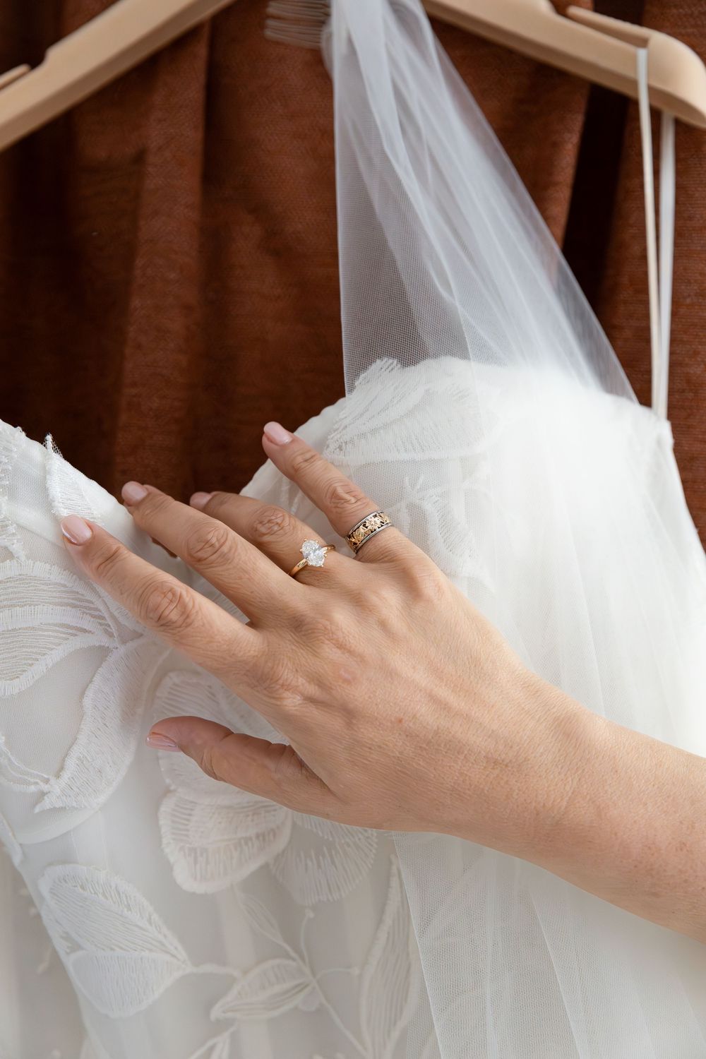 Bride's hand with diamond engagement ring and gold band touching a white lace wedding dress on a hanger in Guelph, Ontario.