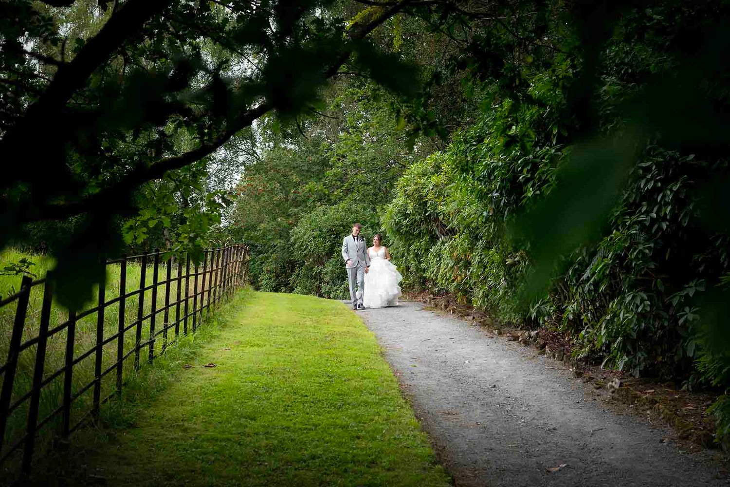 Bride and groom walking hand in hand along a scenic garden path lined with lush greenery and wooden fence.