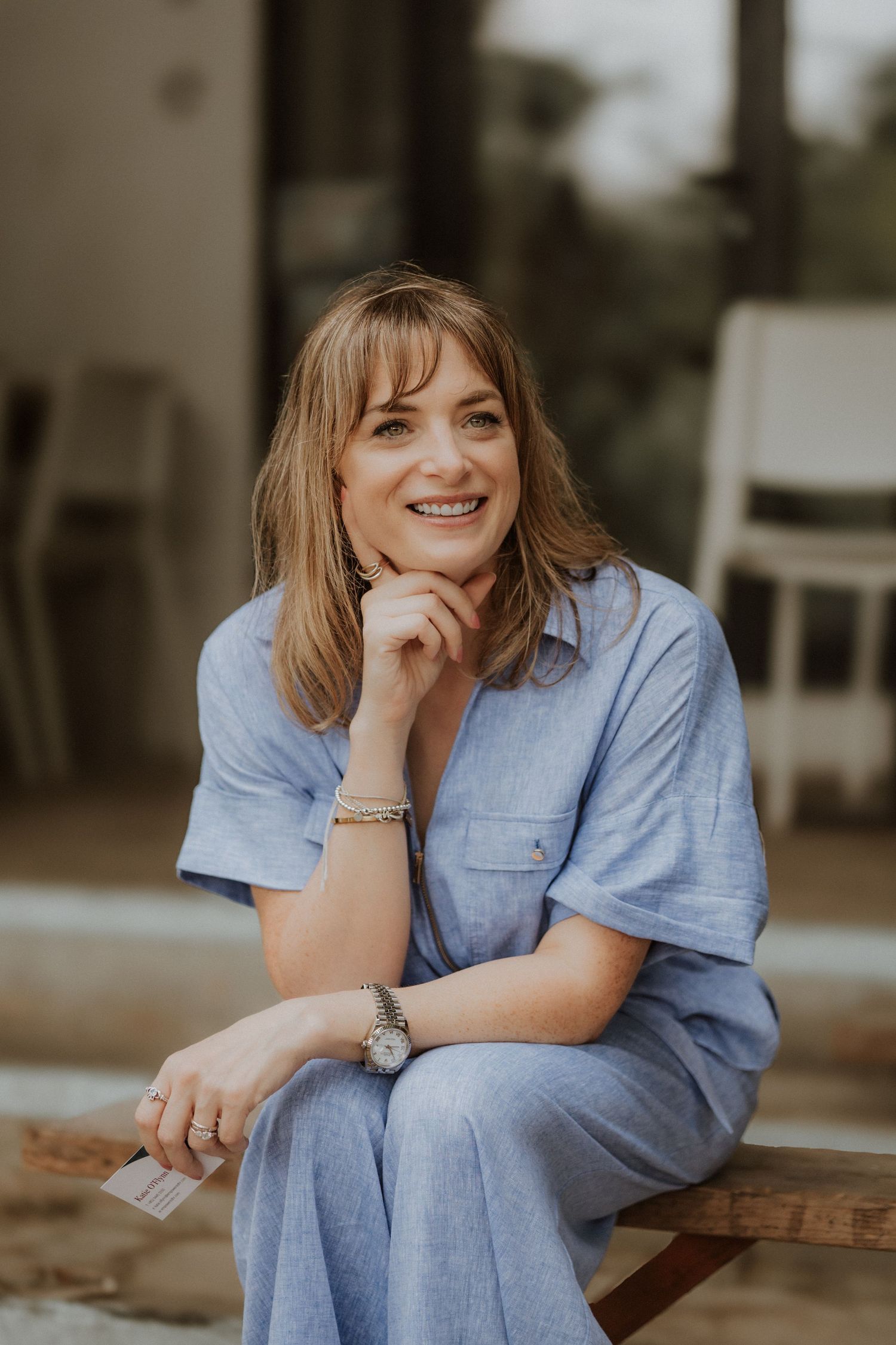 Smiling woman in blue dress sitting outdoors, resting chin on hand, wearing a watch and bracelet.