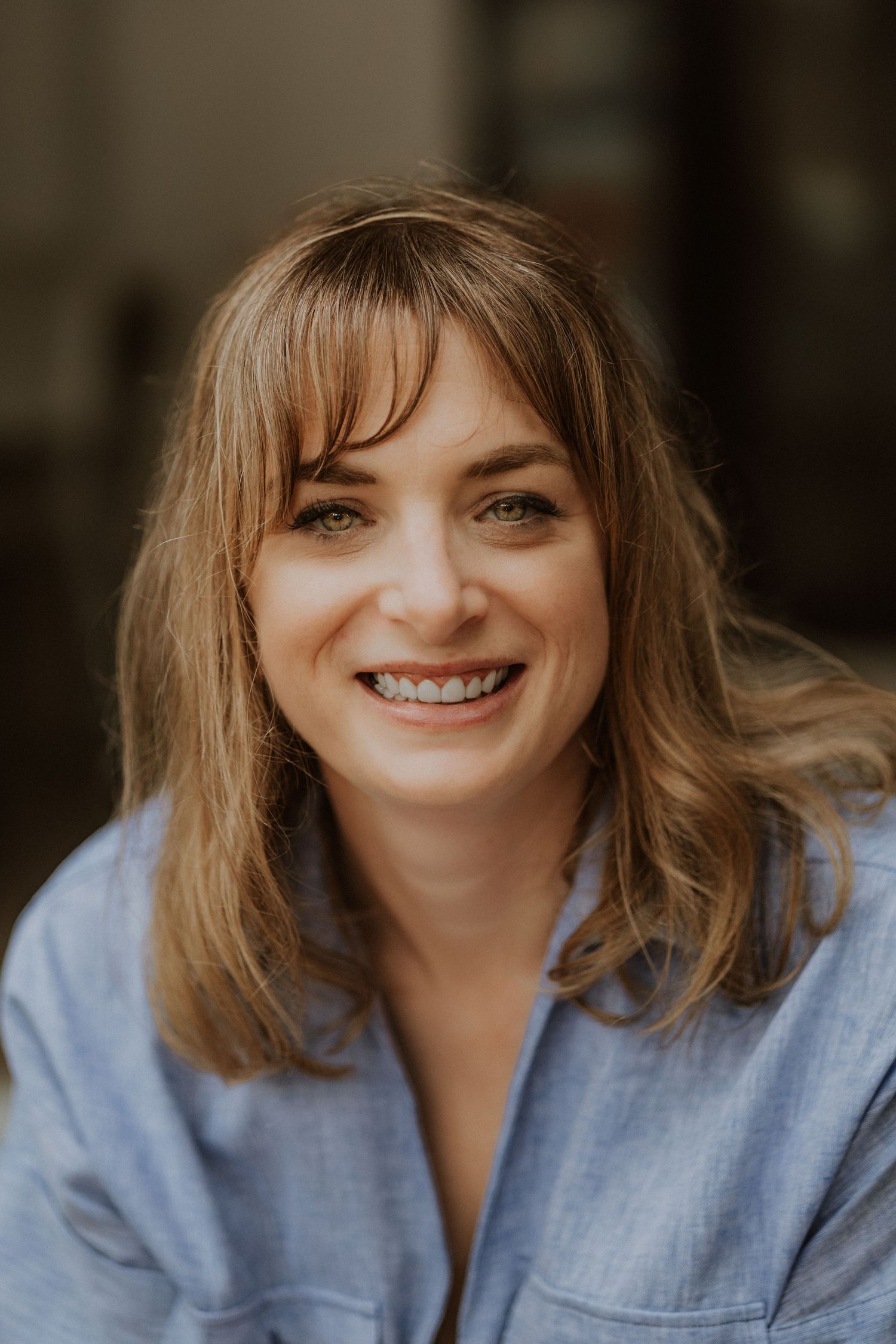 Smiling woman with bangs and shoulder-length brown hair wearing a light blue shirt, photographed outdoors.