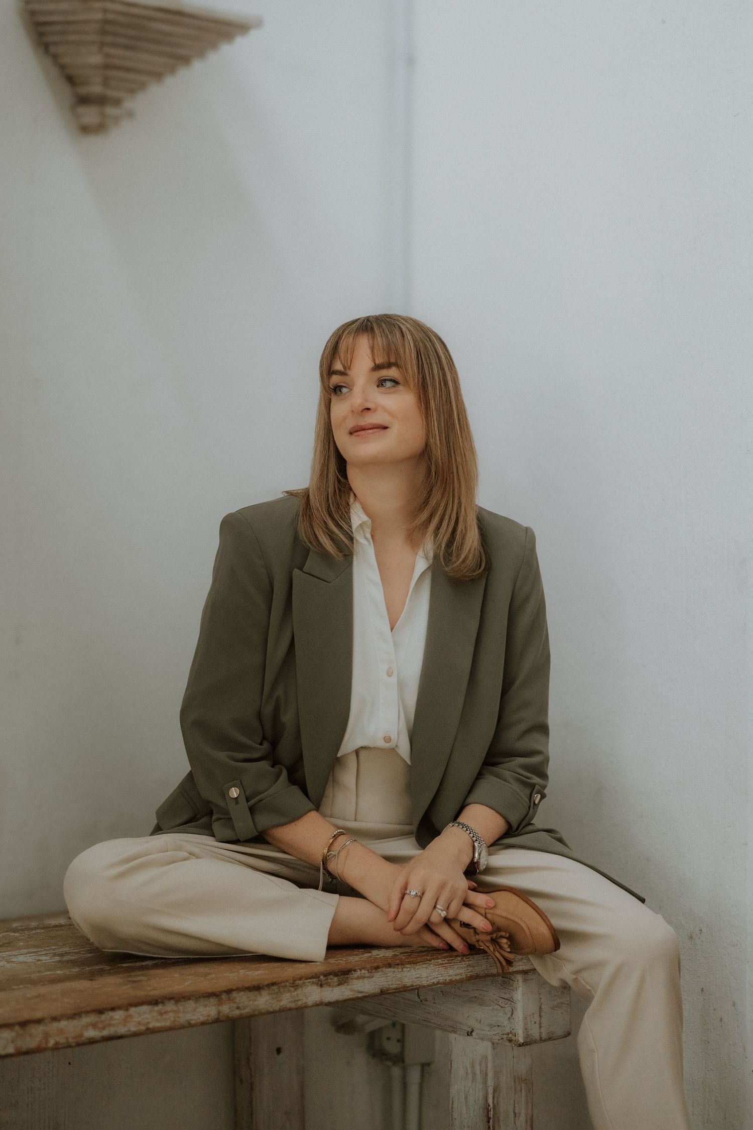 Woman in olive blazer and beige pants sitting cross-legged on a bench, gazing upward in a minimalist white setting.