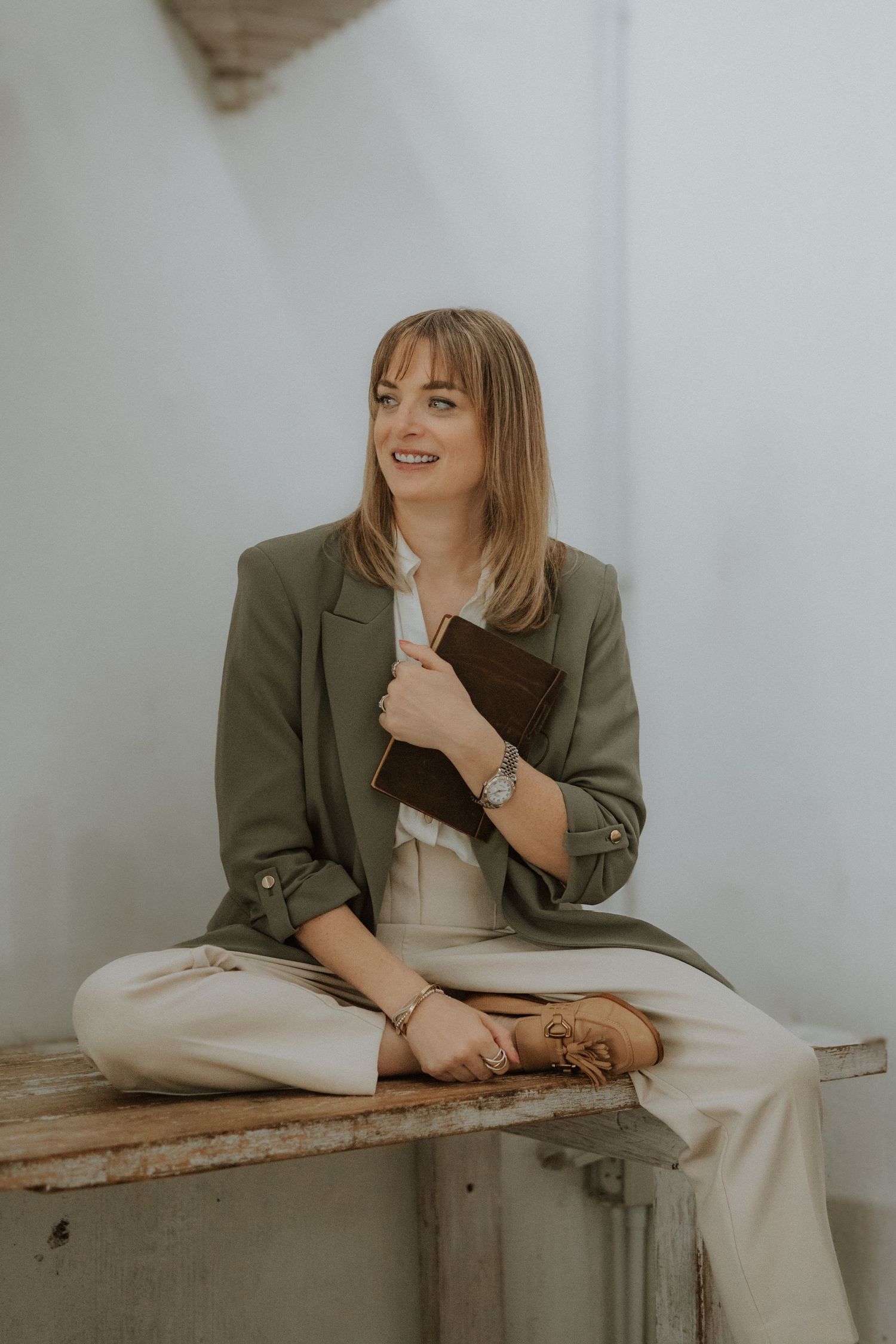 Smiling woman in green blazer and beige pants sitting cross-legged, holding a brown notebook, wearing a watch and bracelets.