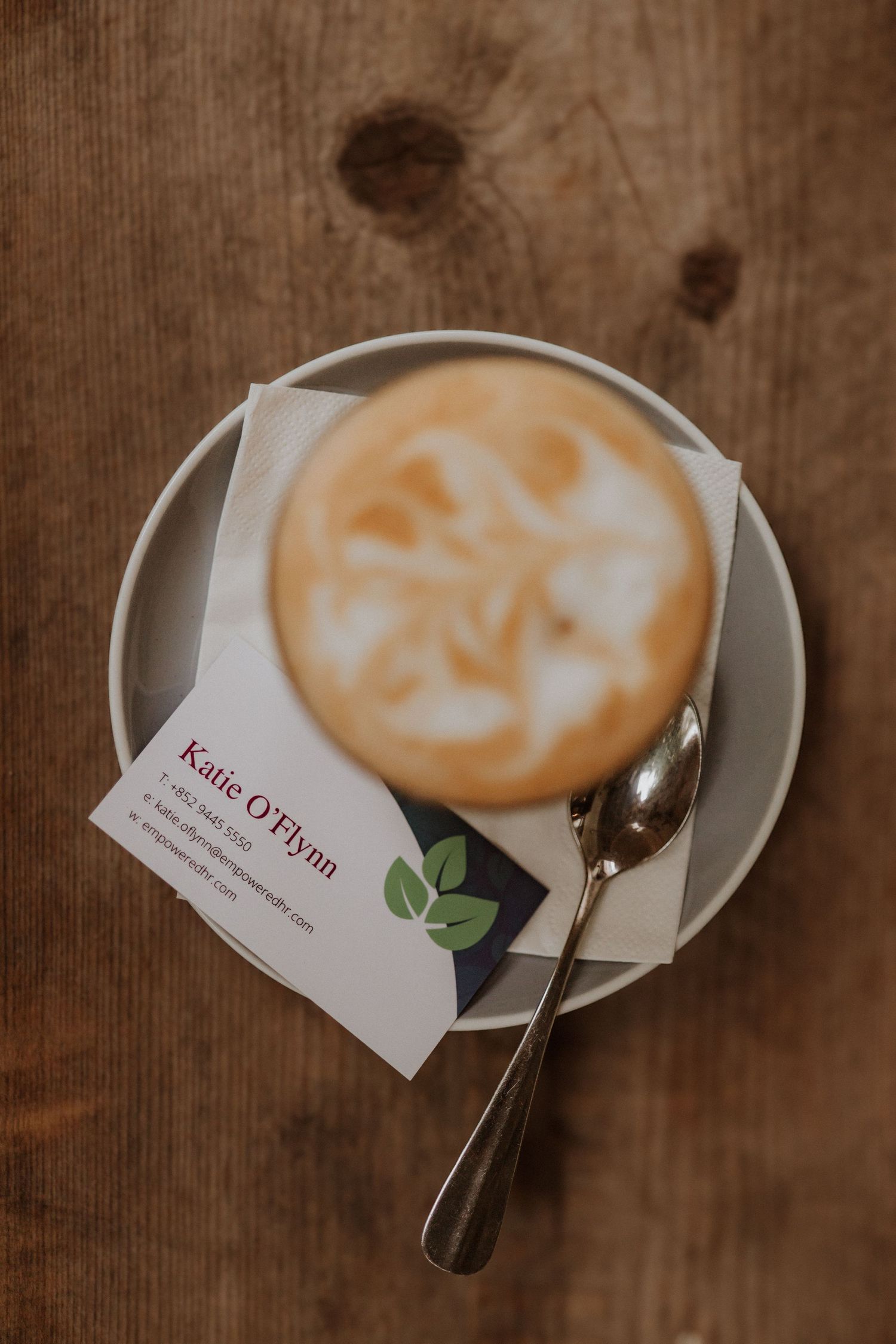 Overhead view of a frothy cappuccino on a saucer with a spoon and Katie O'Flynn business card on a wooden table.
