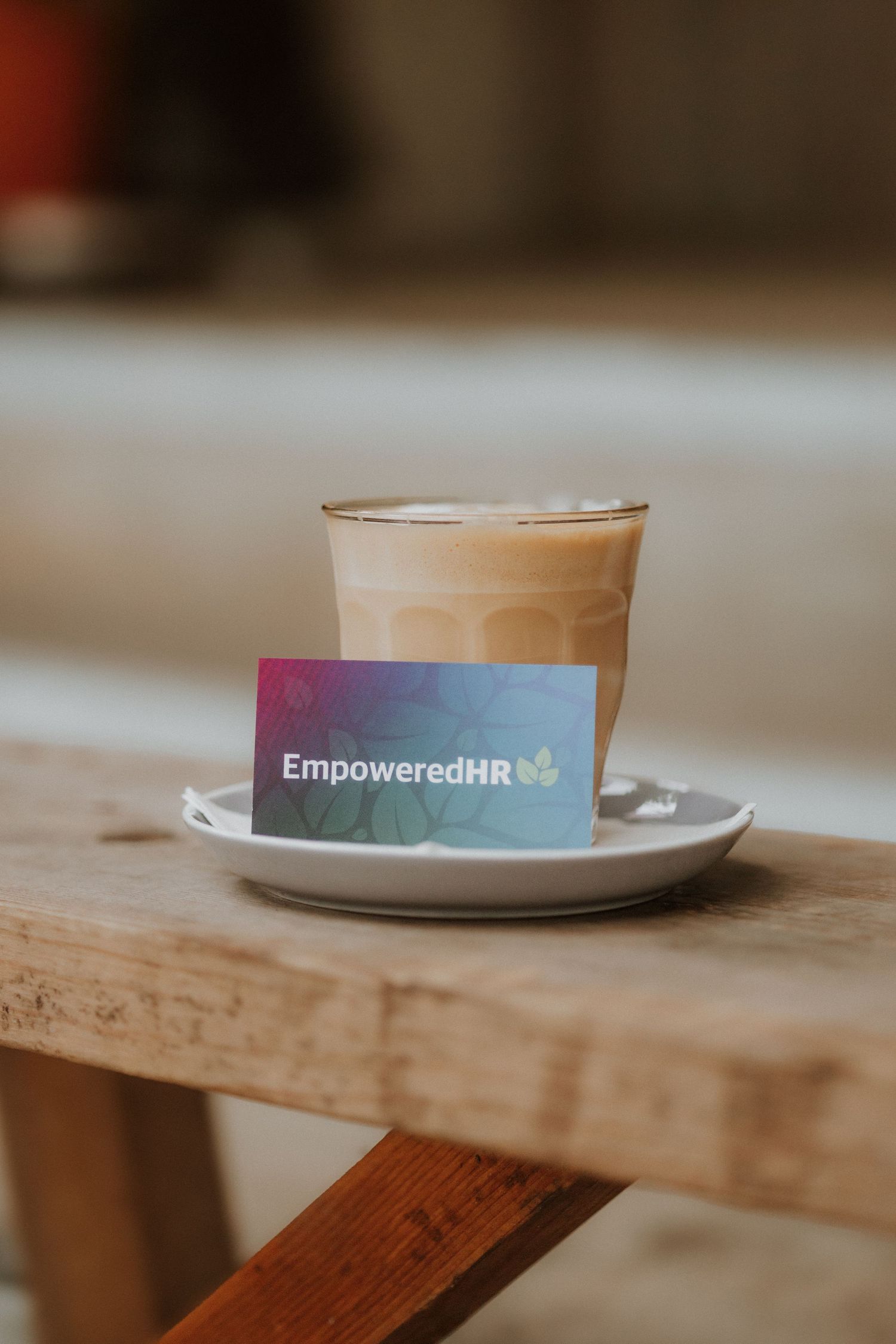 EmpoweredHR business card leaning against a latte coffee cup on a white saucer atop a rustic wooden table.