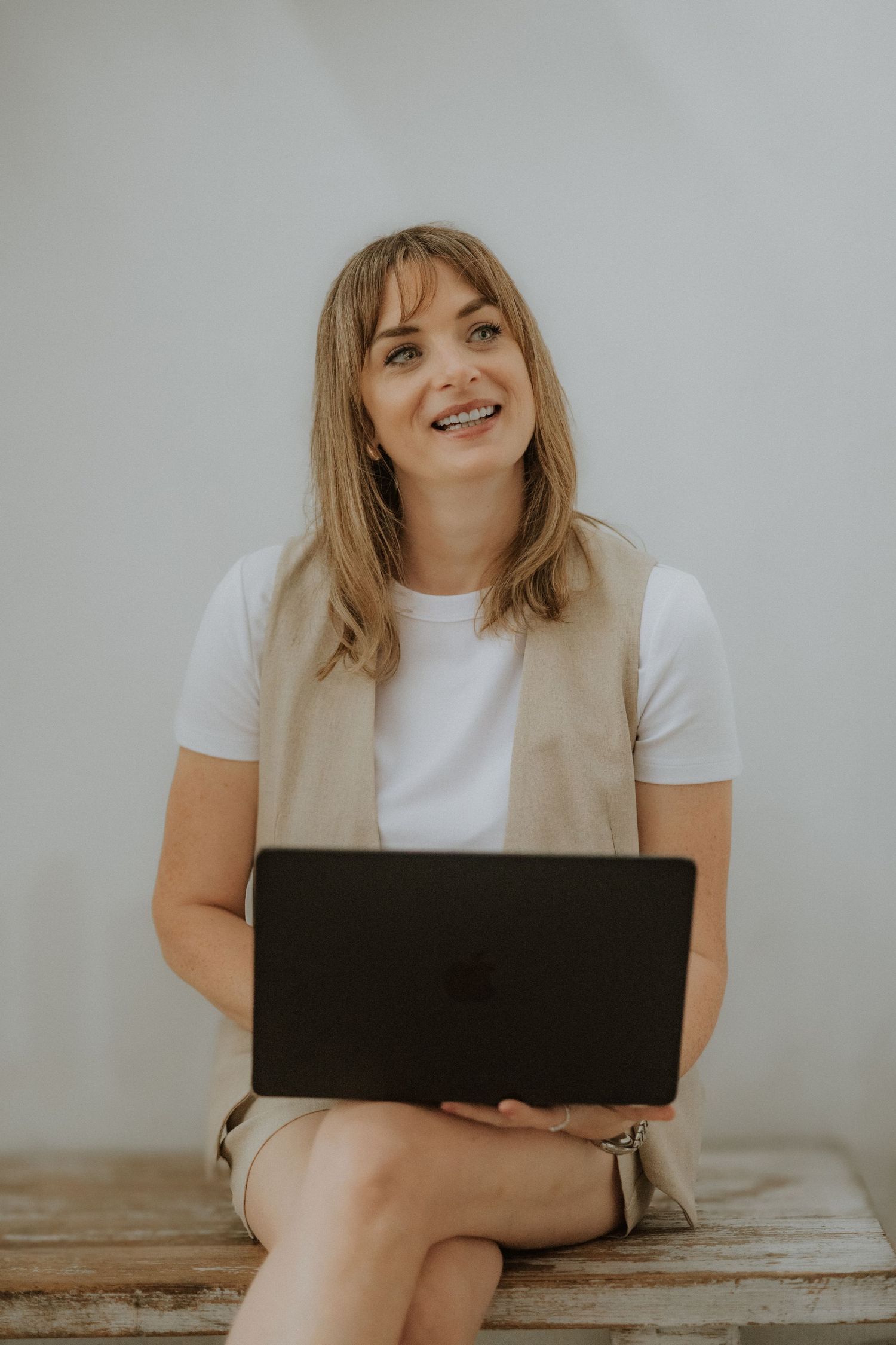 Young woman with braces smiling while holding a MacBook laptop, wearing a white tee and beige vest, seated casually.