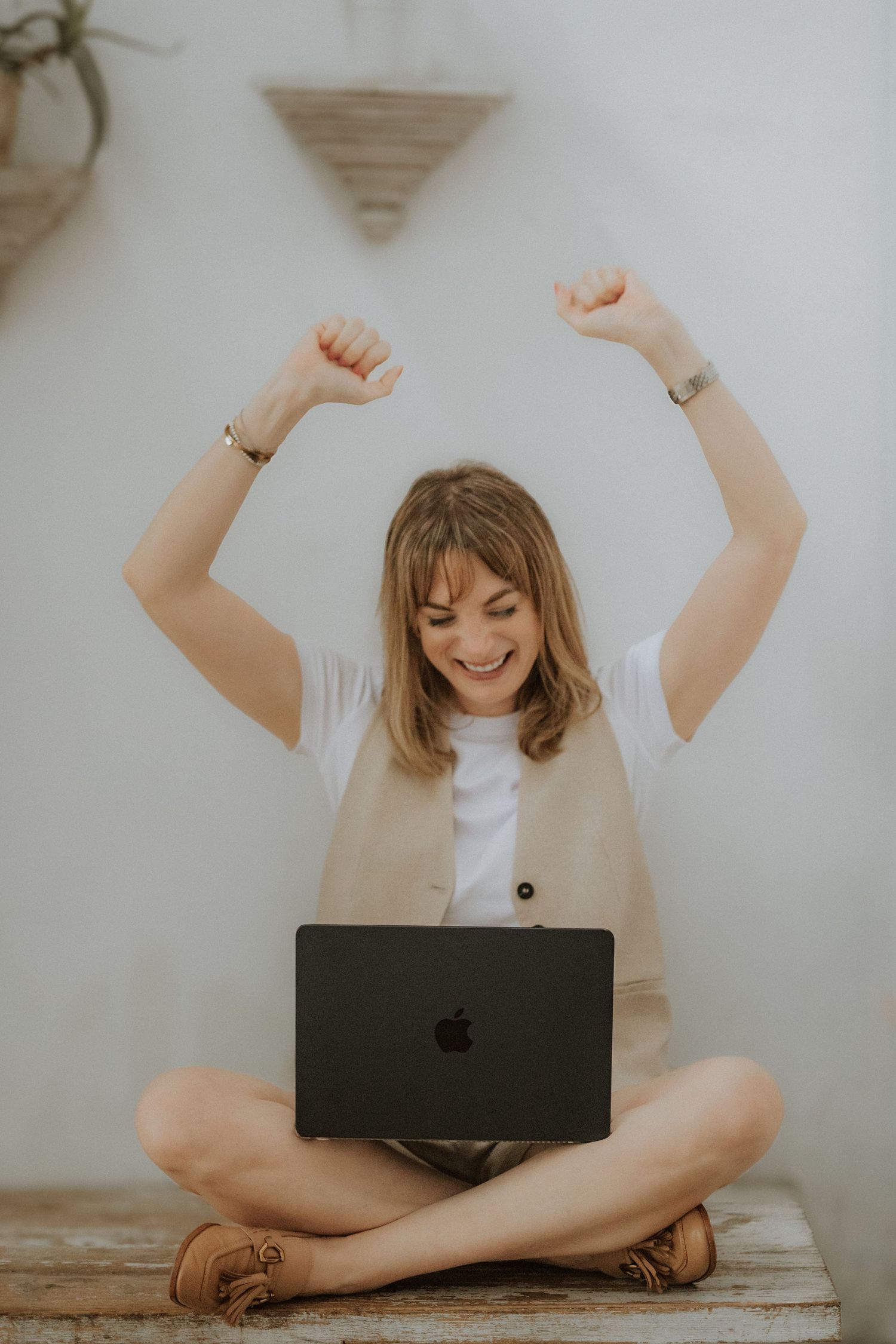 Woman sitting cross-legged with Apple laptop, arms raised in celebration, wearing white tee and beige vest.