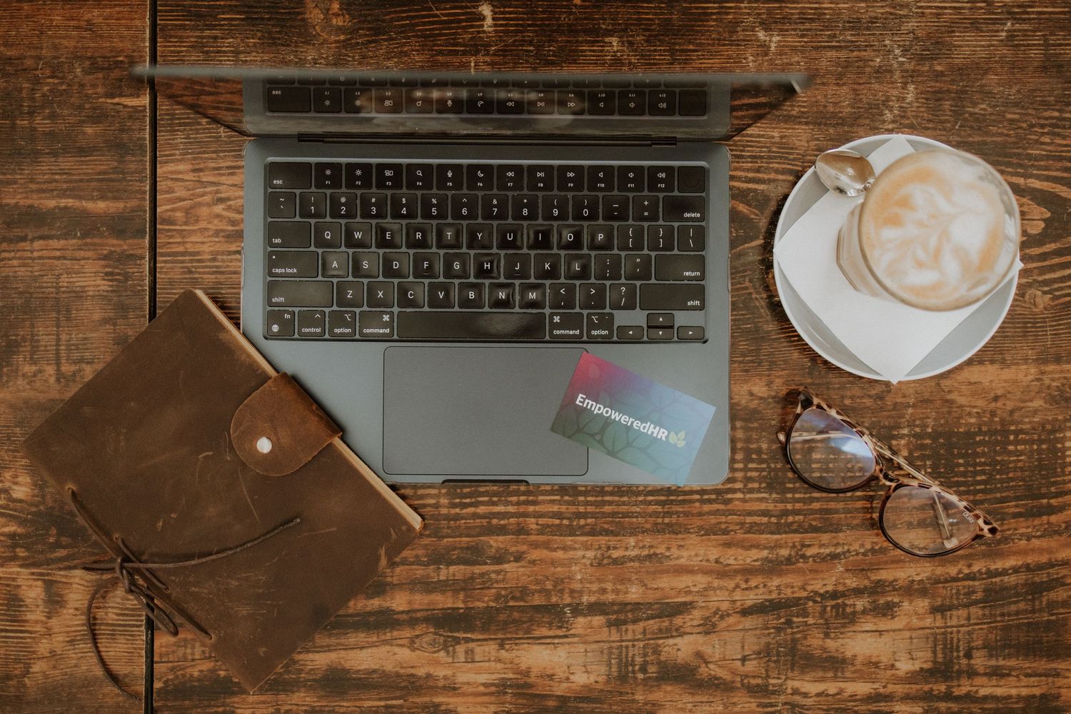 Overhead view of laptop, leather journal, coffee latte, glasses on rustic wooden desk workspace.