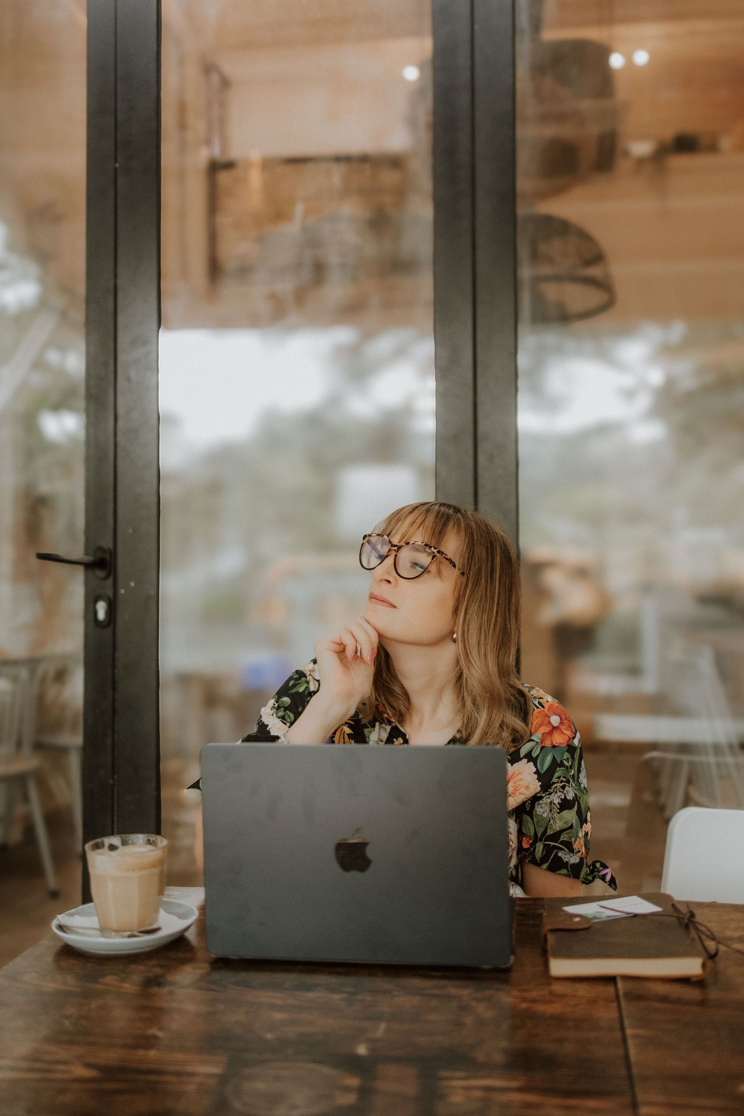 Woman with glasses sitting at wooden table with MacBook laptop and iced coffee, gazing thoughtfully in a cafe.