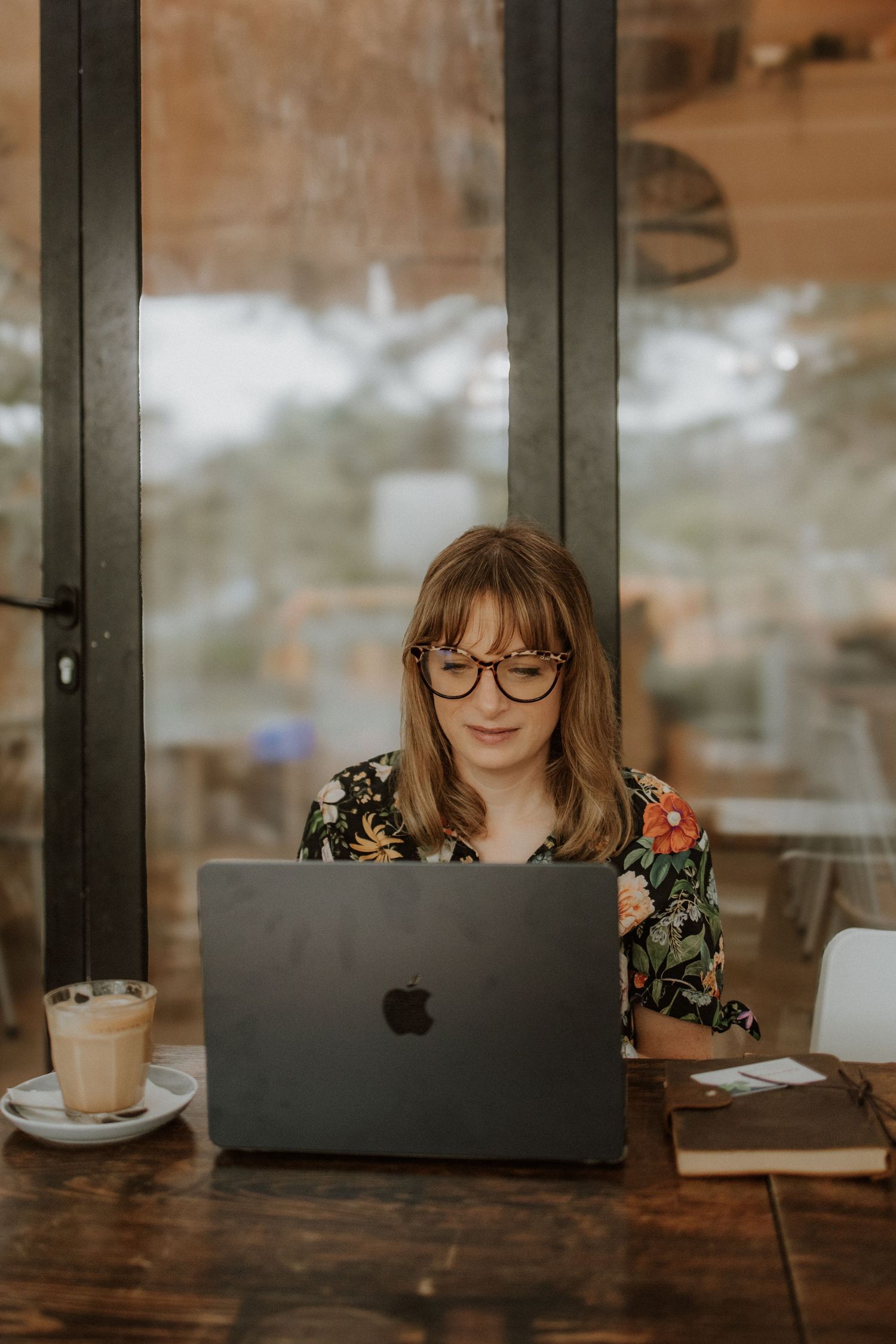 Woman with glasses working on MacBook laptop at wooden cafe table with latte coffee beside her.