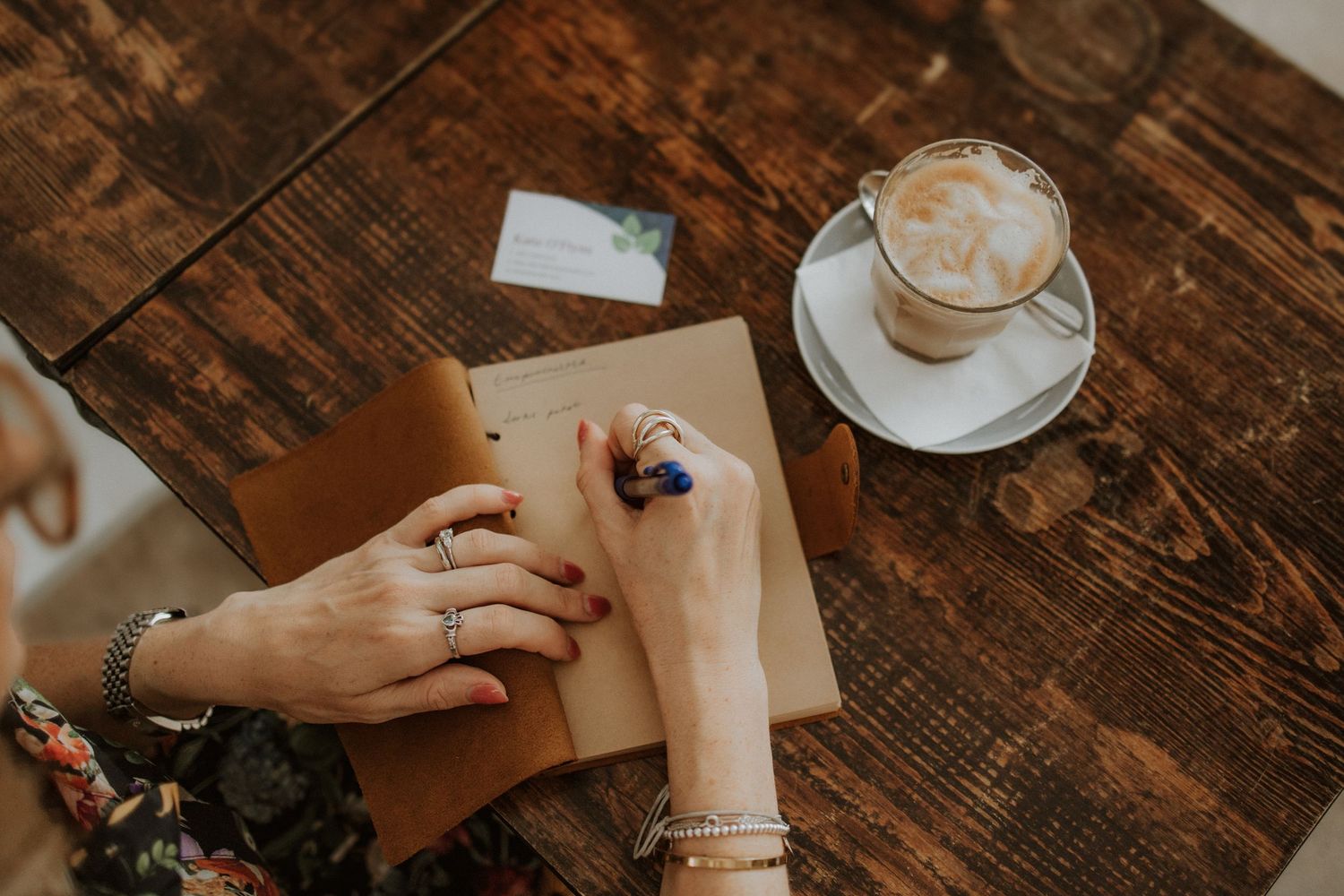 Woman writing in a notebook at a rustic wooden table beside a frothy cappuccino coffee cup.