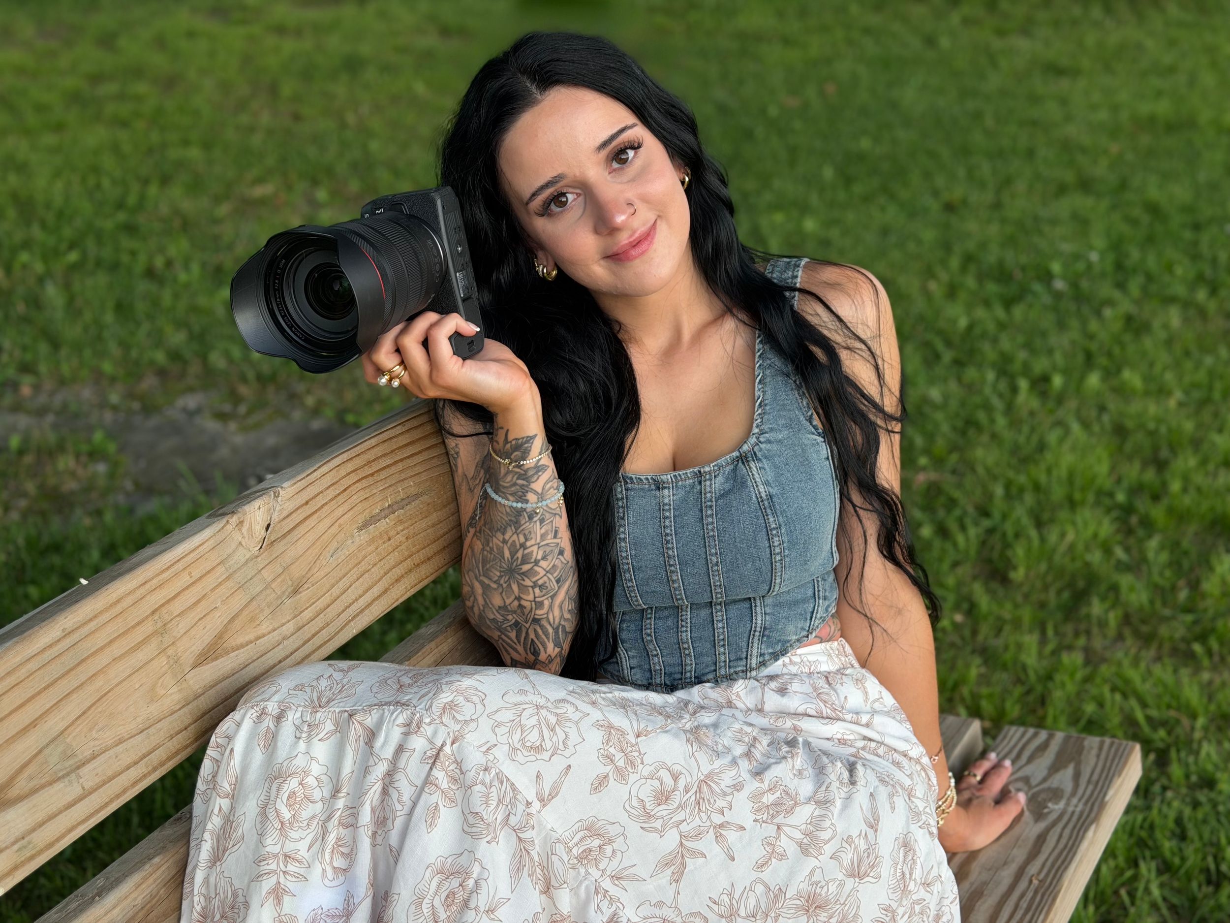 A tattooed woman with long dark hair holds a camera on a wooden bench outdoors, wearing a denim top and floral skirt.