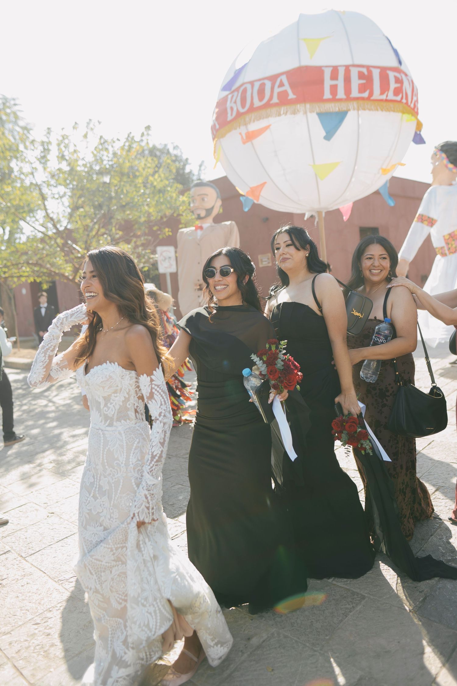 Bride in lace gown walks with bridesmaids in black dresses at outdoor wedding celebration with colorful balloon decor.