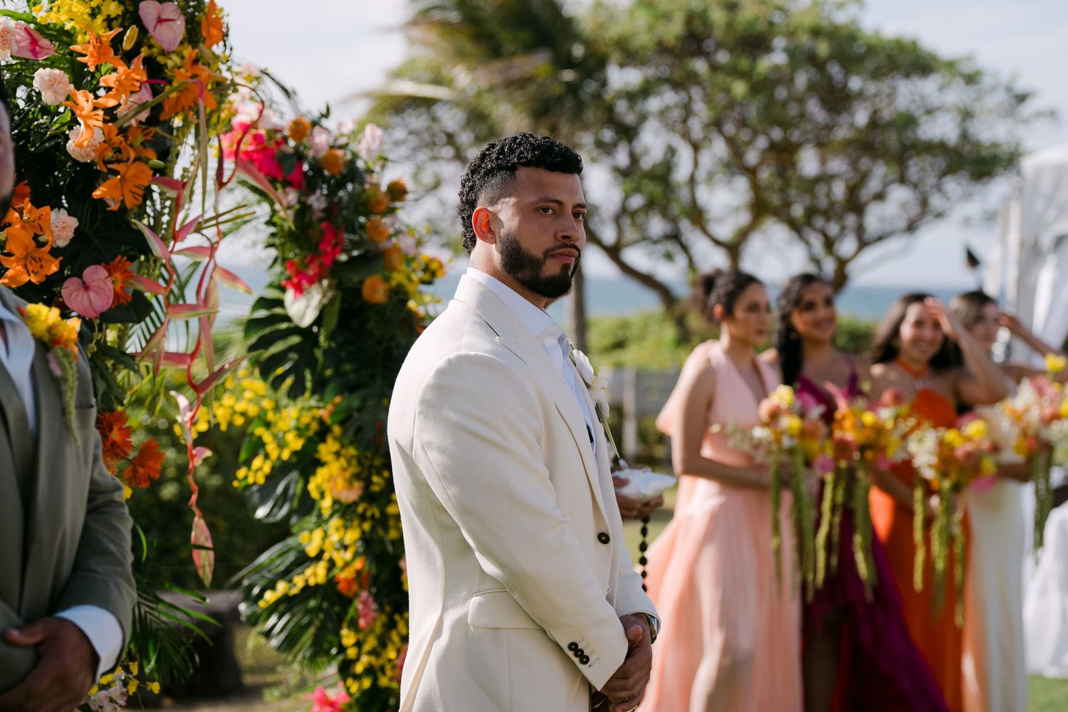 Groom in white suit stands at outdoor tropical wedding altar with colorful floral arch and bridesmaids in background.