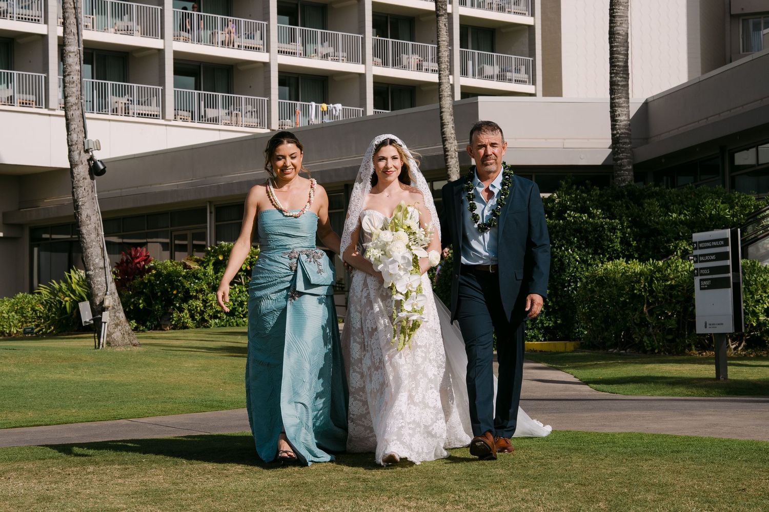 Bride in white lace gown walks with parents on green lawn outside hotel in Hawaii.