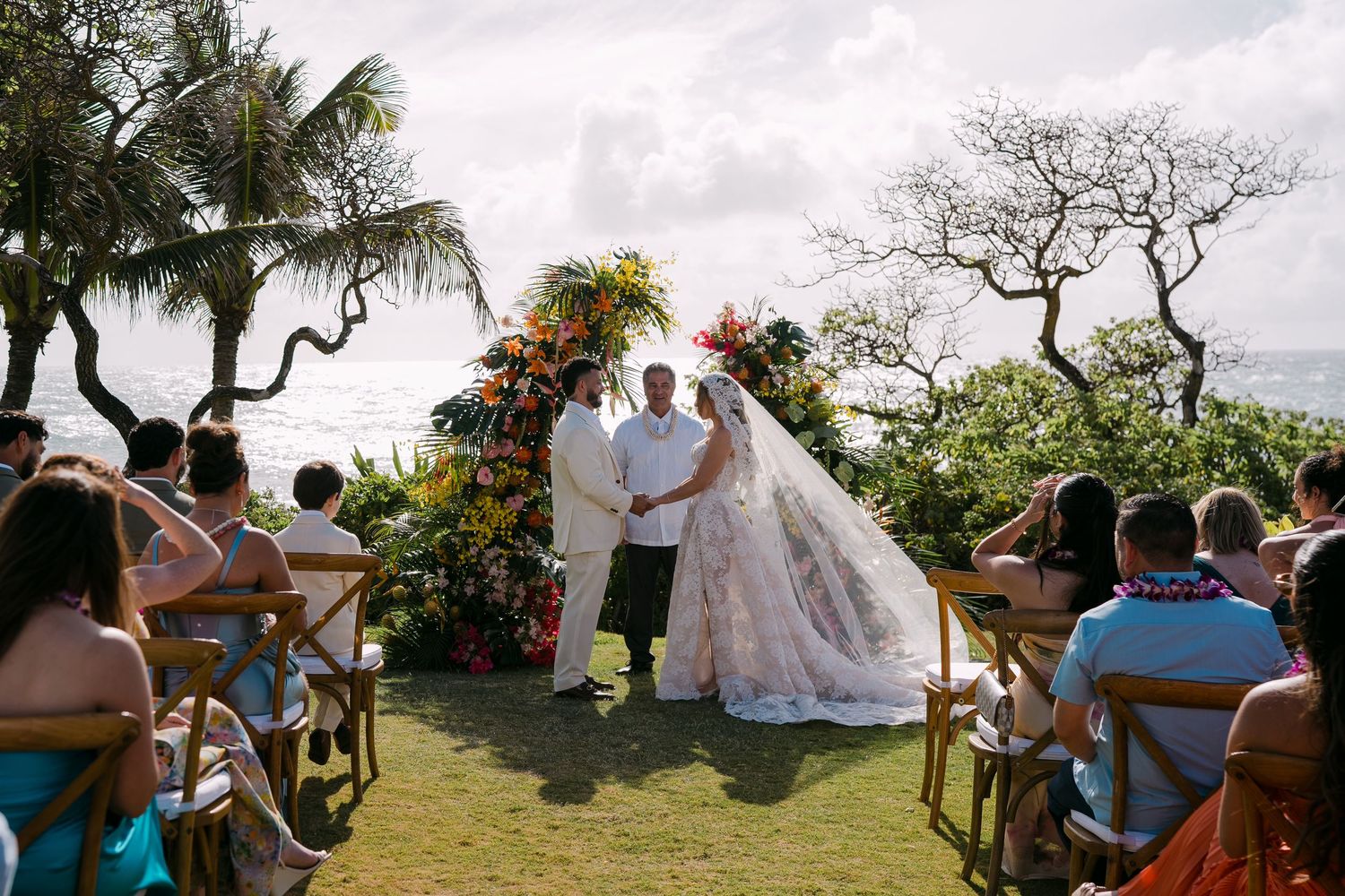 Outdoor beach wedding ceremony with bride and groom at floral arch, guests seated on wooden chairs overlooking ocean.