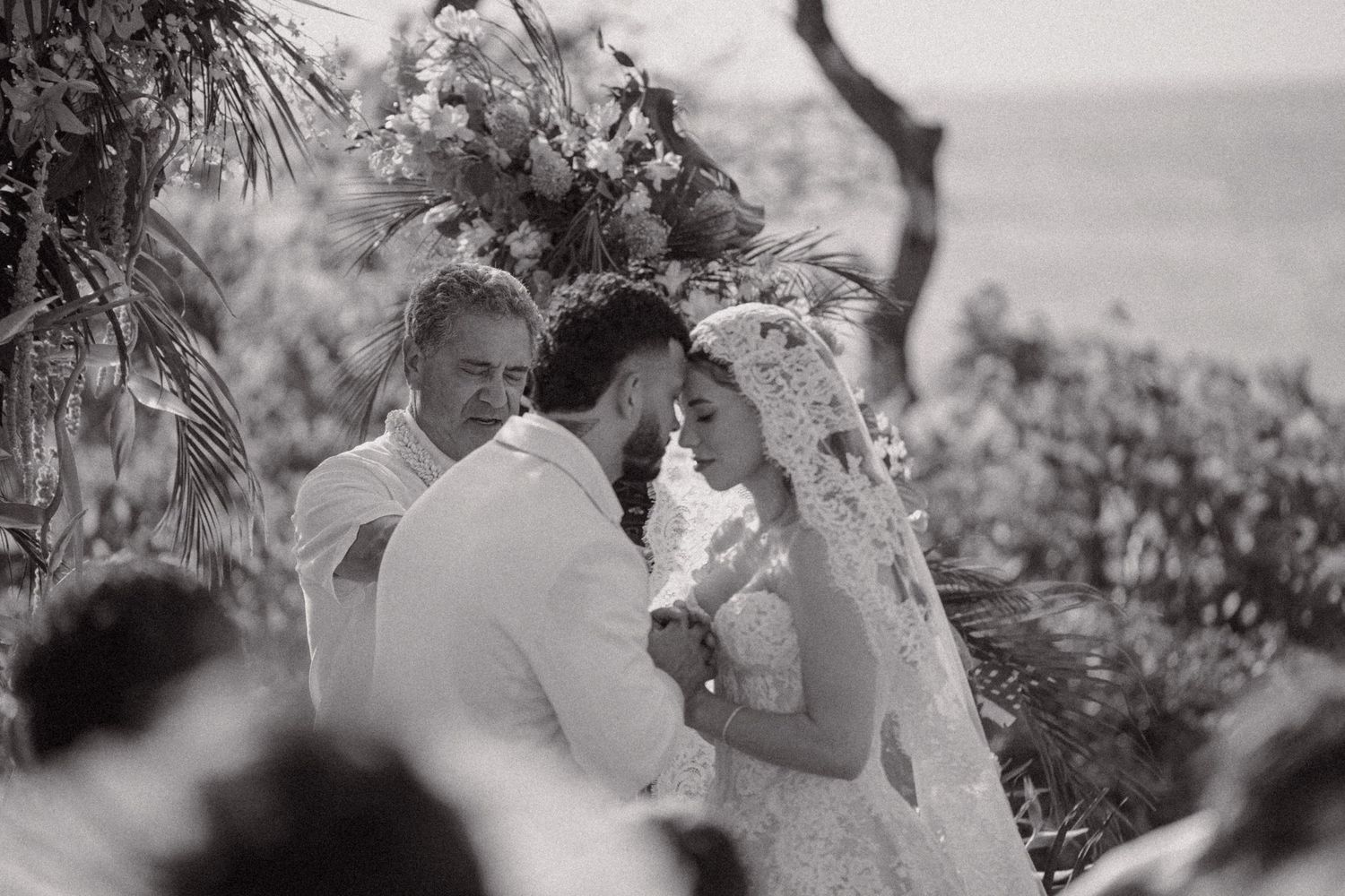 Black and white wedding ceremony photo of bride and groom exchanging vows outdoors with floral arch backdrop.