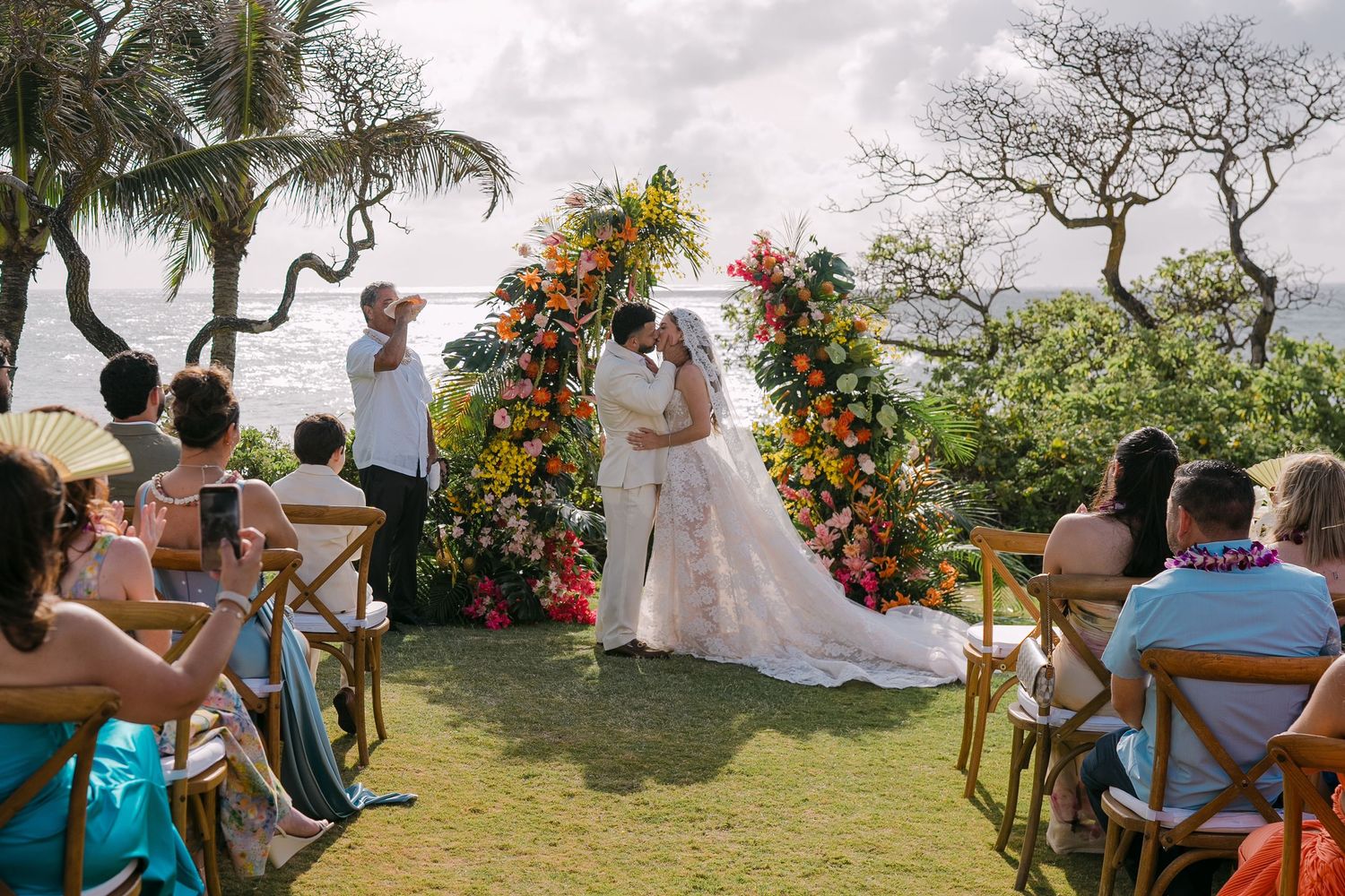 Couple kissing at outdoor tropical wedding ceremony with colorful floral arch, palm trees, and ocean view in Maui.