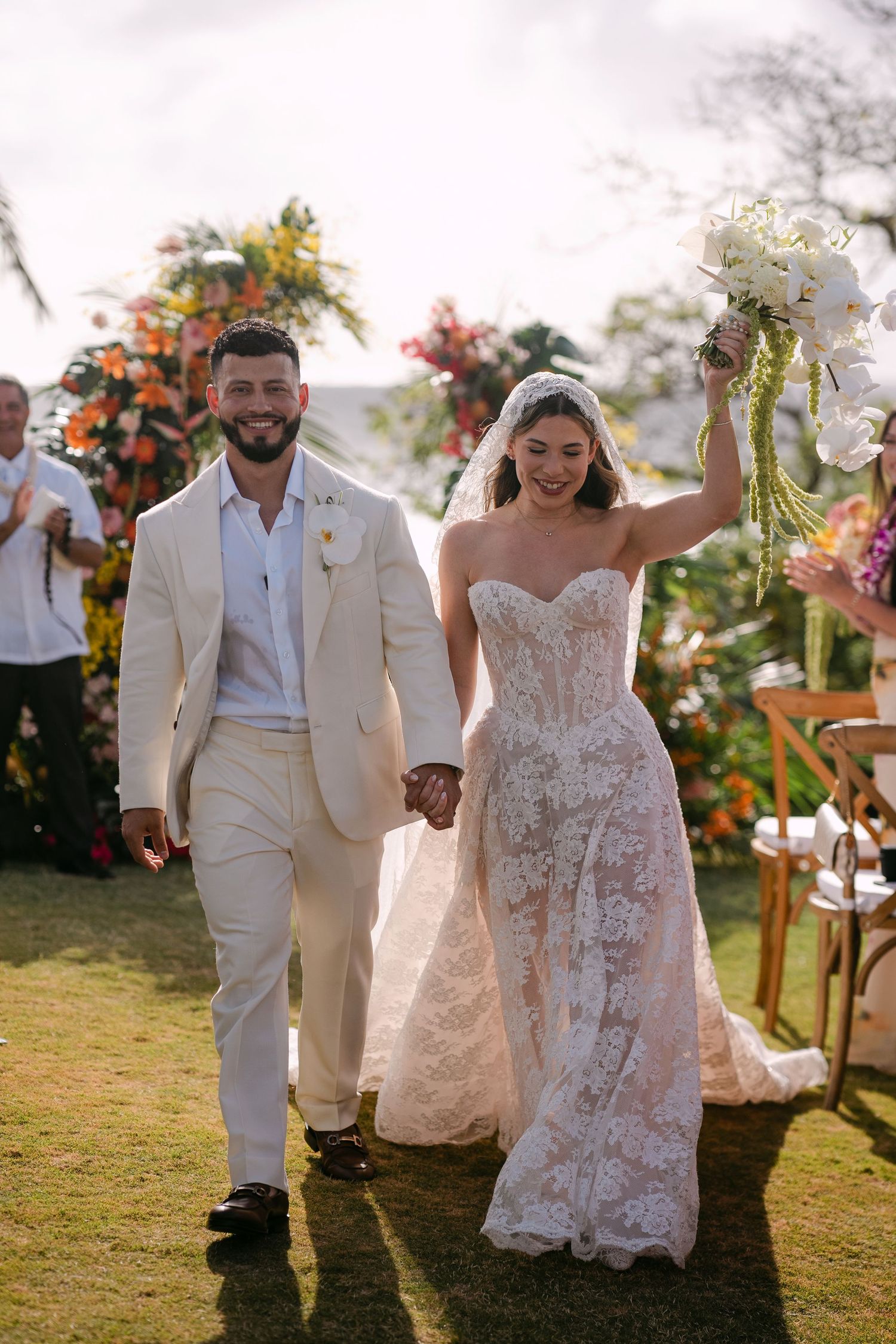 Joyful newlyweds walk down the aisle, bride in lace gown holding bouquet aloft, groom in cream suit, outdoor garden wedding.