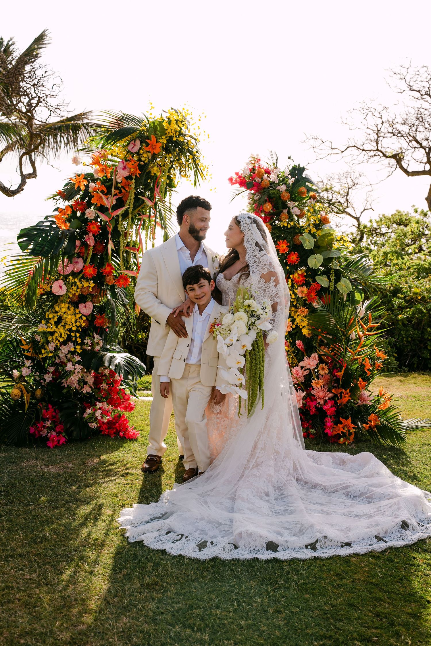 Bride and groom with child pose before vibrant tropical floral arch at outdoor wedding ceremony.