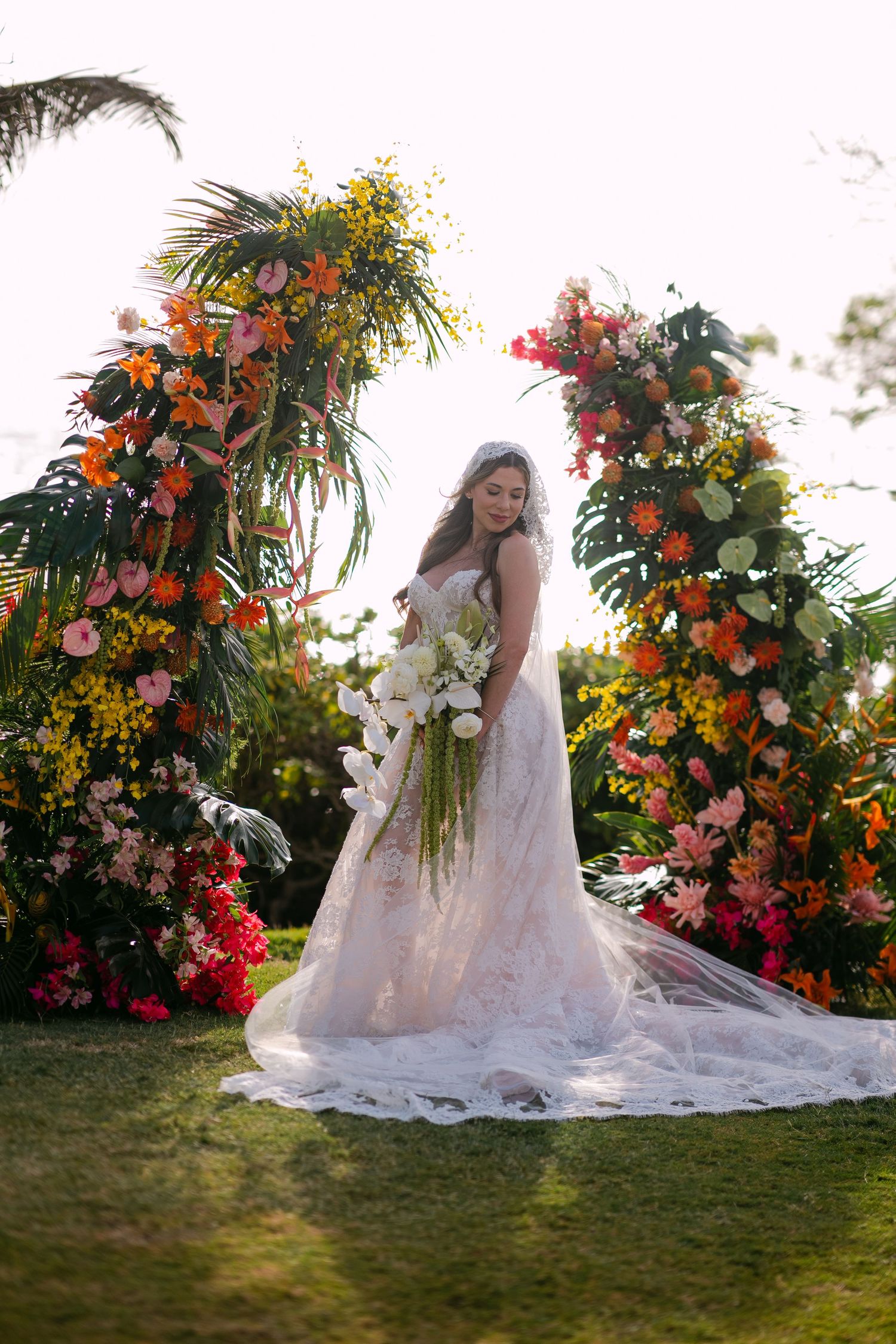 Bride in white lace gown holding floral bouquet stands before vibrant tropical floral arch on green lawn.