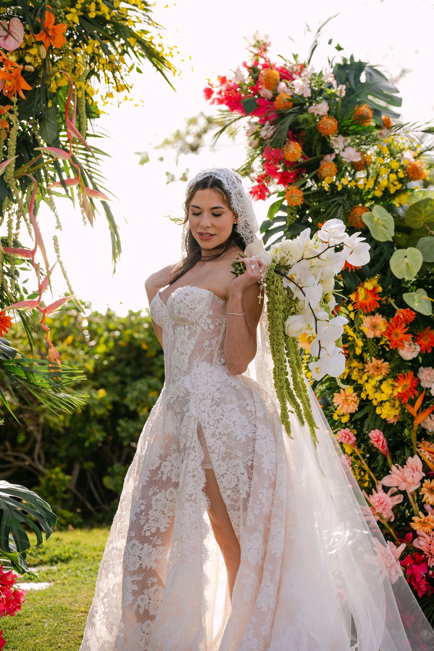 Bride in lace wedding gown with veil poses by colorful floral arch outdoors.