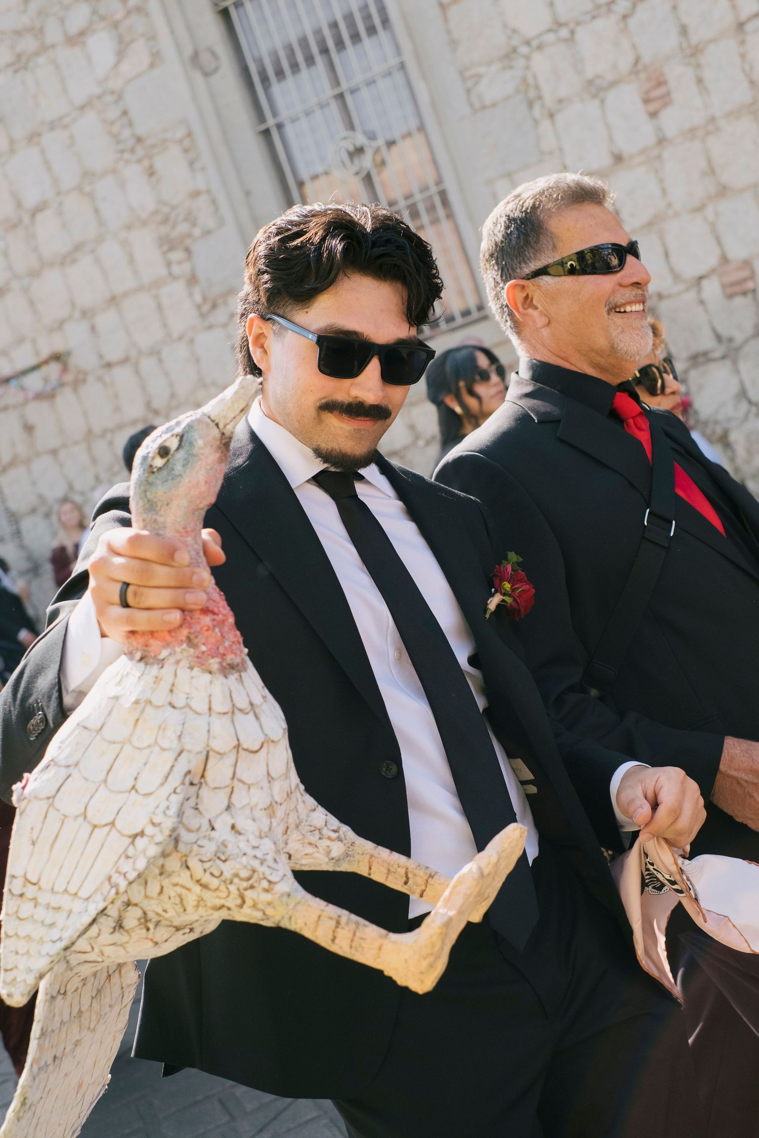 Man in black suit and sunglasses holds a large decorative bird statue at a formal outdoor event.