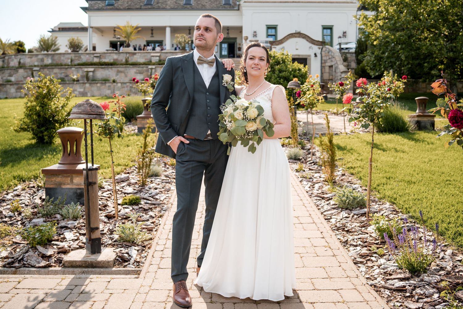 Bride and groom posing in a garden with roses, bride holding a bouquet, elegant venue in the background.