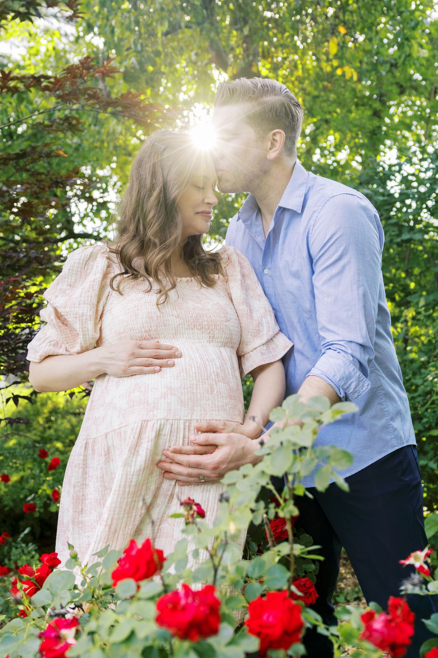 Pregnant couple embracing in a sunlit garden with red roses, man kissing woman's forehead while holding her baby bump.