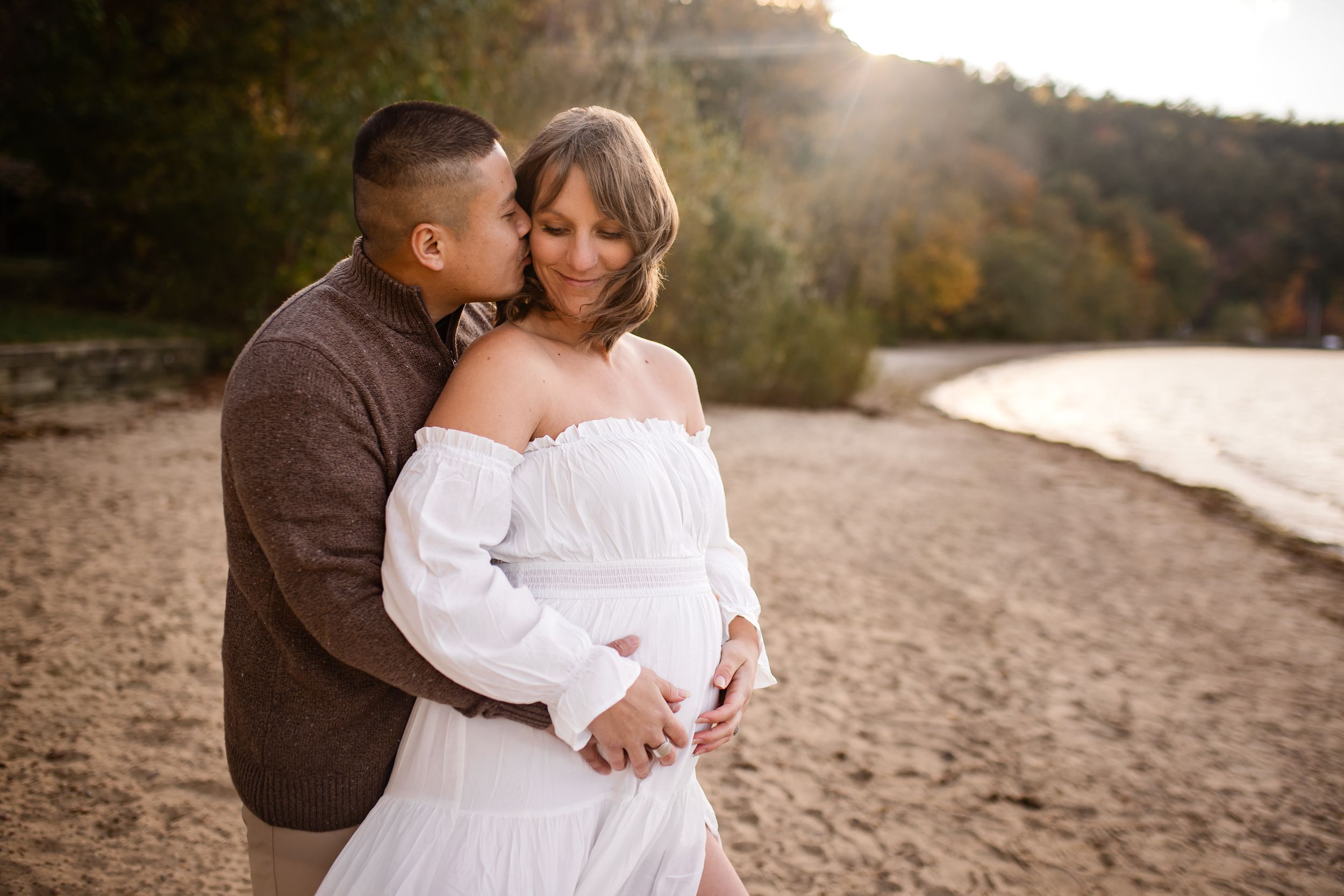 Couple embracing on sandy beach at golden hour, pregnant woman in white off-shoulder gown with partner in brown shirt.