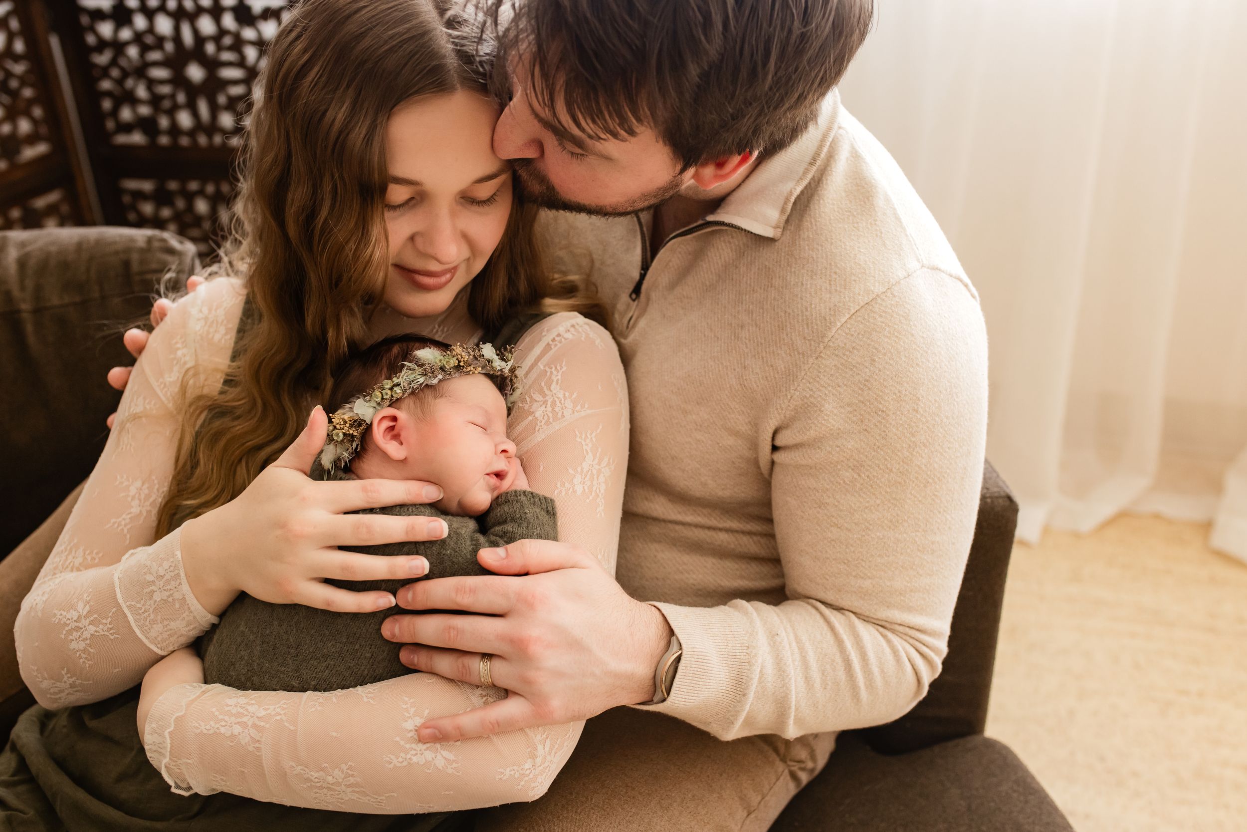 New parents sitting together and kissing their sleeping newborn baby wearing a floral headband. Madison WI Newborn photographer.