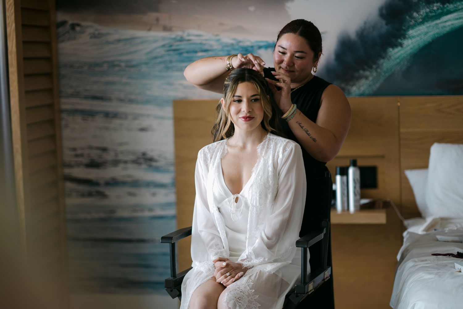 A bride in a white robe laughs joyfully while a makeup artist applies her bridal makeup before the wedding ceremony.