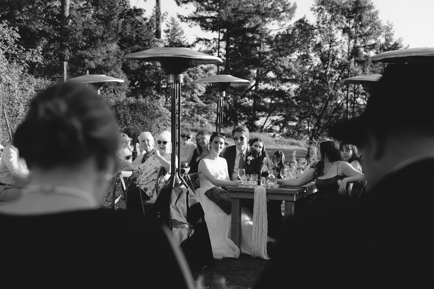 Black and white outdoor wedding reception with guests gathered around tables under patio heaters and umbrellas.