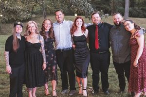 Group of eight adults posing together outdoors at a formal event, dressed in elegant attire on a green lawn.