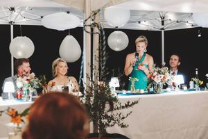 A bridesmaid in green gives a speech at an elegant outdoor wedding reception under a white tent with balloon decor.