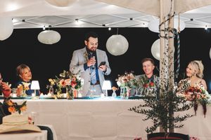 Man giving a wedding speech at a decorated reception table with floral centerpieces under a white tent.