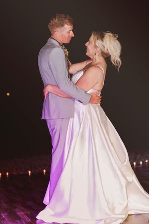 Bride and groom share their first dance, bride in white satin gown, groom in gray suit, illuminated by soft purple lighting.