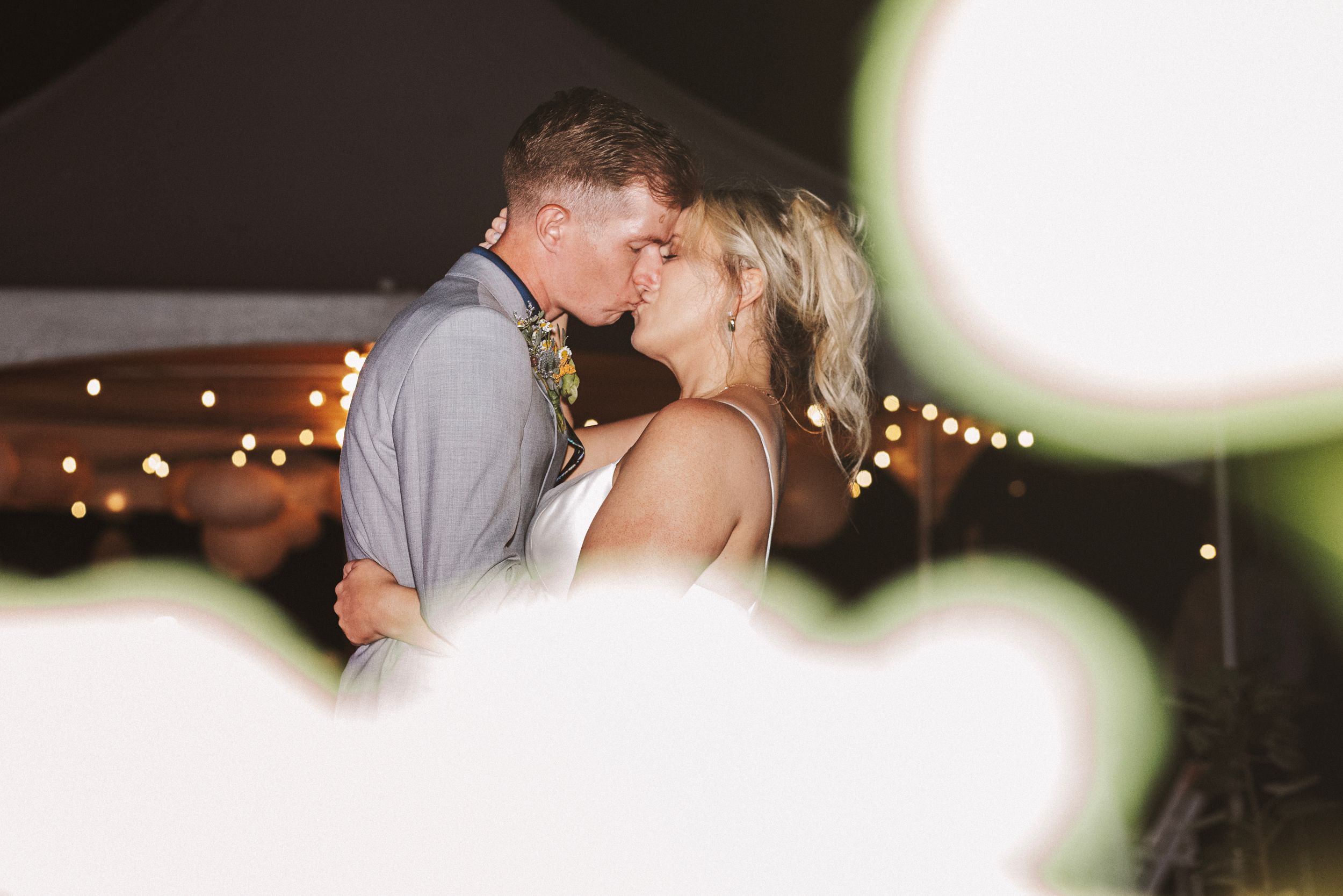 Bride and groom sharing a romantic kiss under twinkling fairy lights at their outdoor wedding reception.