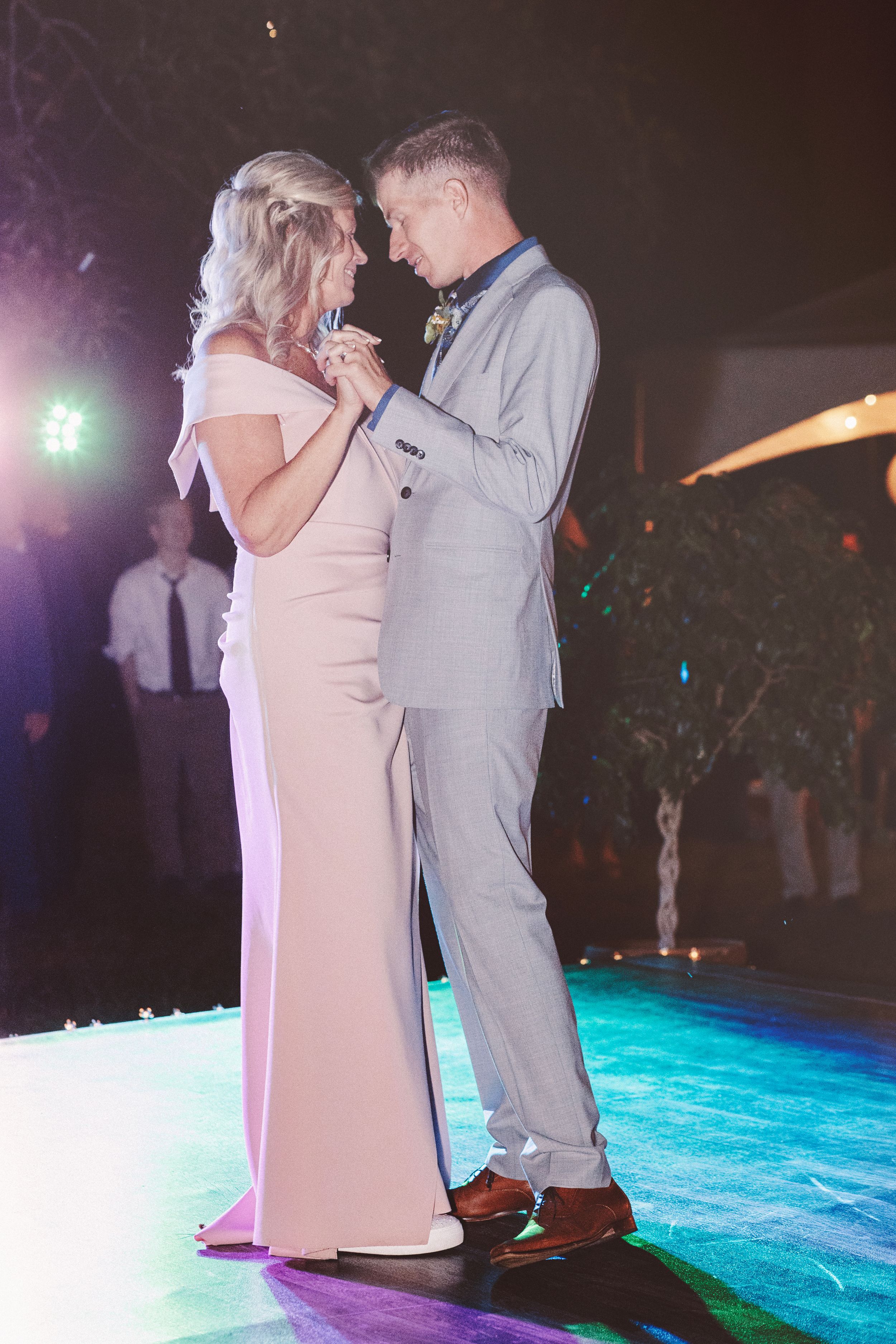 Couple sharing first dance at wedding reception, woman in pink gown and man in gray suit under colorful lights.