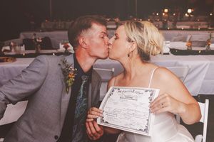 Newlywed couple kissing while holding their marriage certificate at wedding reception.