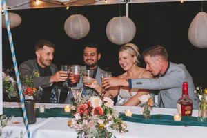 Wedding party toasting with drinks at reception table decorated with flowers and white paper lanterns overhead.