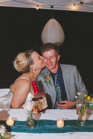 Bride kissing groom on cheek at sweetheart table with candles and whiskey bottle at wedding reception.