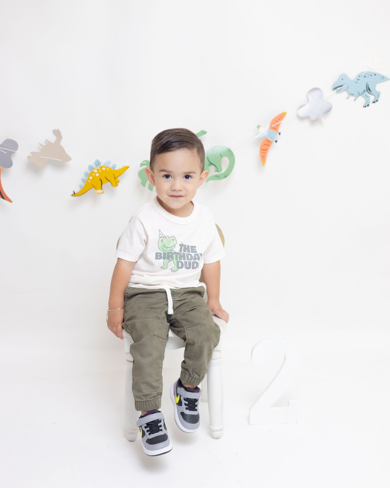 Toddler boy in birthday dino shirt and olive pants sits on white stool with colorful dinosaur decorations behind him.